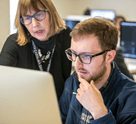 Male student working at the computer with professor looking over shoulder offering help