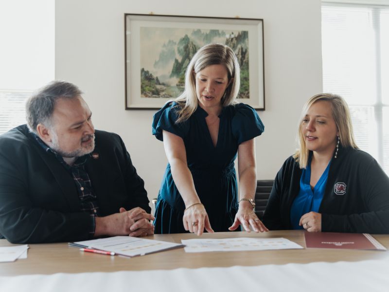 Male and two women are looking over papers on a table. 