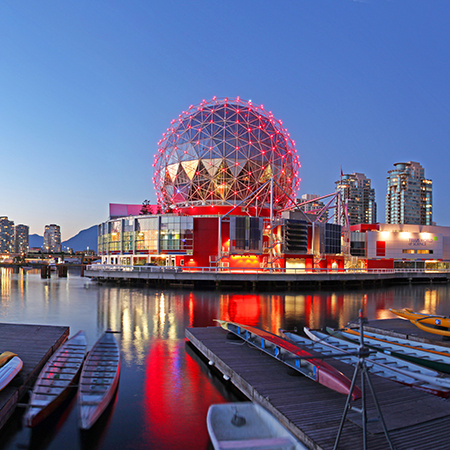 The illuminated geodesic dome of Science World reflects on the water at dusk in Vancouver, with colorful boats in the foreground.