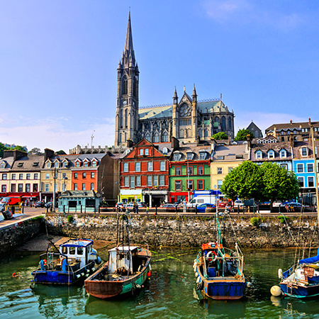 Colorful waterfront houses and fishing boats line the harbor in Cobh, Ireland, with the tall spire of St. Colman’s Cathedral rising above the town under a clear blue sky.