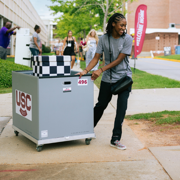 student in gray shirt pulling gray box of belongings on sidewalk during move-in